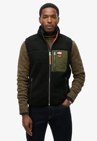 Sort fleece vest med olivengrøn brystlomme, lynlås lukning og badge detaljer. Båret over en struktureret brun sweater og mørkeblå jeans.