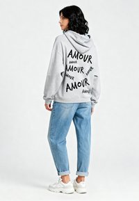 Felpa grigia con cappuccio e stampa nera "AMOUR" sul retro, abbinata a jeans in denim blu chiaro e sneakers bianche.