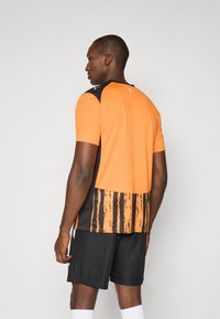 Orange atletisk skjorte med sorte accenter, prydet med et struktureret stribemønster på ryggen. Parret med sorte shorts og hvide sokker.