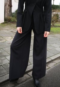 Melns pielāgots žakets ar pieguļošu siluetu un divām pogām, apvienots ar platām melnām biksēm, abiem izgatavotiem no gludas auduma.