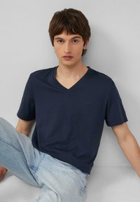 T-shirt blu navy con scollo a V realizzato in morbido cotone, caratterizzato da maniche corte e un sottile dettaglio del logo, abbinato a jeans azzurri chiari.
