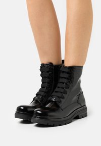 Bottes courtes en cuir verni noir avec bouts ronds, surface texturée, devant à lacets, talon renforcé et semelles en caoutchouc antidérapantes.