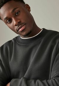 Sudadera negra con un cuello redondo acanalado, que presenta una textura suave y un corte holgado. Los acentos incluyen un detalle blanco en el cuello interior.