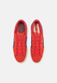 Røde ruskindsneakers med blå accenter, tekstureret tunge, gummisål og flade snørebånd; logo synligt på overdelen og sålen.