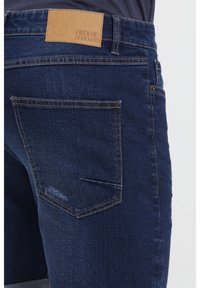 Tamnoplave traperice s kožnim umetkom na struku, dvostrukim šavovima i prepoznatljivim dizajnom stražnjeg džepa.