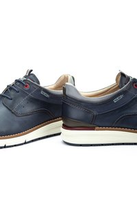 Zapatos de cuero azul marino con detalles perforados, suela de goma color tostado y acentos de tejido gris. Presenta cordones azules y ojales rojos.