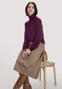 Violetter Rollkragenpullover mit lockeren Ärmeln, kombiniert mit einem beigefarbenen, knöchellangen Rock und braunen knielangen Stiefeln, sitzend auf einem Holzstuhl.