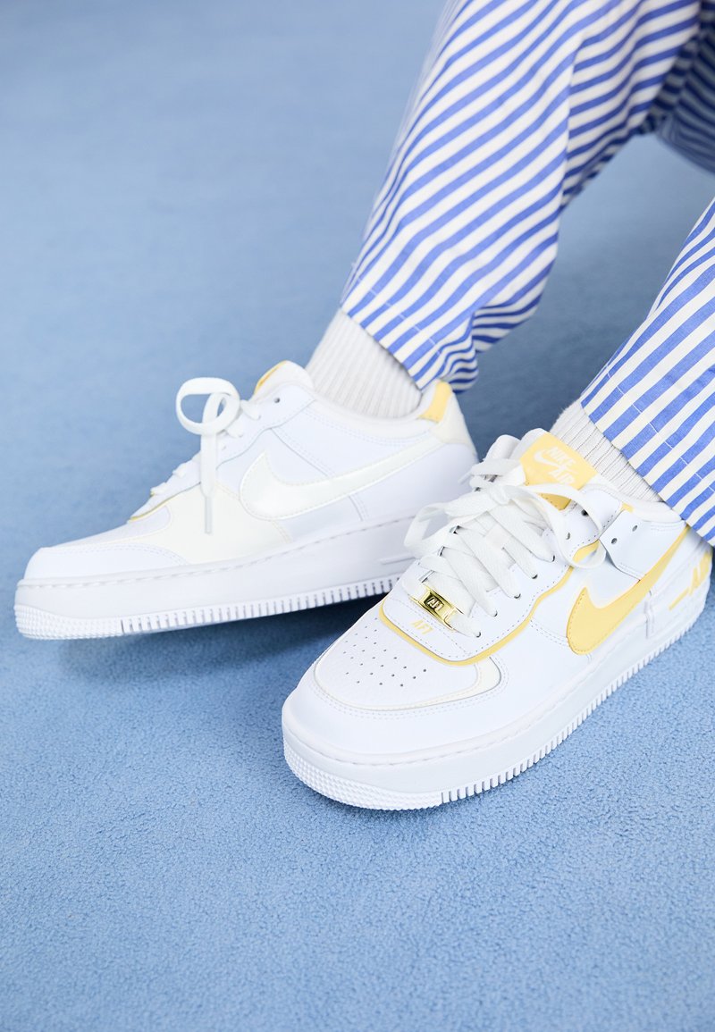 Bijele Nike Air Force 1 tenisice s žutim akcentima i perforiranim prednjim dijelom, uparene s prugastim plavim i bijelim hlačama. Teksturirana plava pozadina.