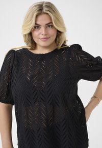 Black blouse with leaf-patterned cutouts, short puff sleeves, and a round neckline. Made of textured fabric with a loose fit.
