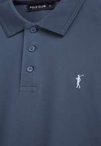 Polo blu con tre bottoni, logo del golfista bianco sul petto e etichetta "Polo Club" sul colletto che indica la taglia media.