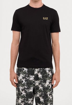 Mężczyzna w czarnej koszulce EA7 Emporio Armani oraz czarnych, białych i szarych pikselowych szortach moro na jednolitym tle.