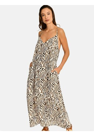 Femme portant une robe maxi sans manches, ample, avec un imprimé zèbre abstrait marron et blanc, posant avec les mains dans les poches sur un fond uni.