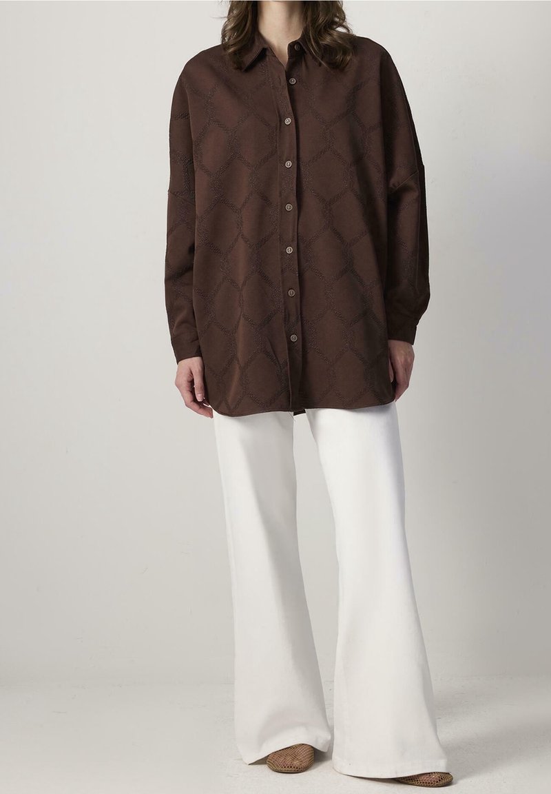 Personne portant une chemise ample marron à motifs avec boutons, manches longues, un pantalon blanc à jambes larges et des chaussures marron tissées.