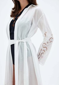 Biały przezroczysty kimono z długimi rękawami, związane w talii. Posiada misternie wycięte wzory wzdłuż brzegów rękawów. Lekki materiał.