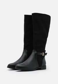 Bottes noires montantes jusqu'au genou, combinant cuir et suède, avec une fermeture éclair latérale, une semelle plate et une petite sangle à accent métallique.