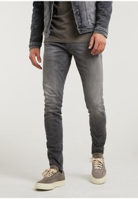 Pantalones vaqueros de mezclilla gris con un ajuste slim, que presentan un acabado deslavado, detalles cosidos y bolsillos frontales, combinados con zapatillas marrones y una chaqueta gris.