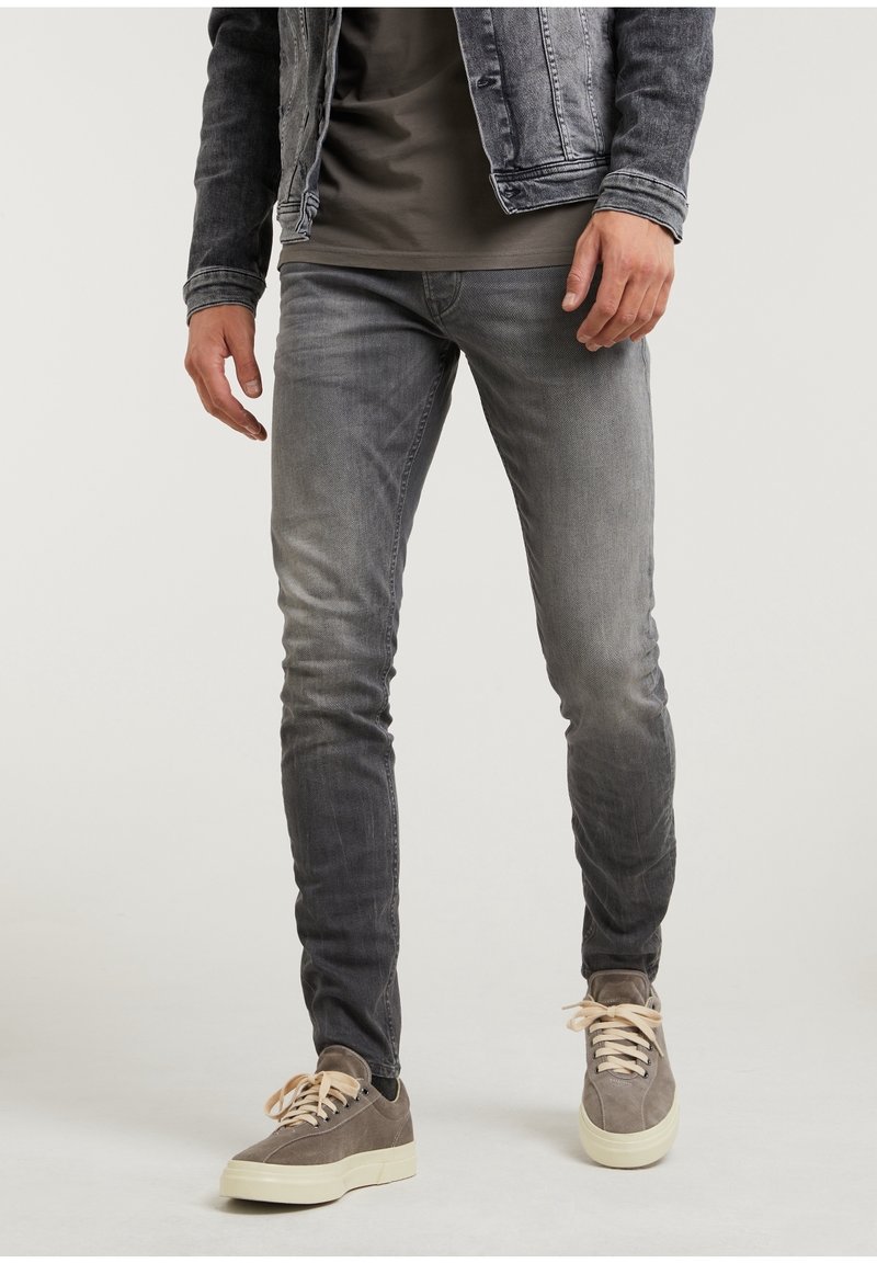 Pantalones vaqueros de mezclilla gris con un ajuste slim, que presentan un acabado deslavado, detalles cosidos y bolsillos frontales, combinados con zapatillas marrones y una chaqueta gris.