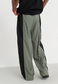 Sports pants featuring a black and olive green design, with white side stripes and a loose fit. Made from a smooth, lightweight material.