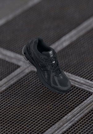 Zwarte sportieve schoen met een bovenwerk van mesh, getextureerde accenten en een gewatteerde zool. Vetersluiting en logo-details aan de zijkant.