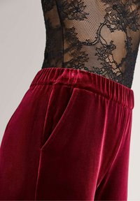 Close-up van een zwarte bloemenkant top, gecombineerd met rode fluwelen broeken met een elastische tailleband en een zijzak.