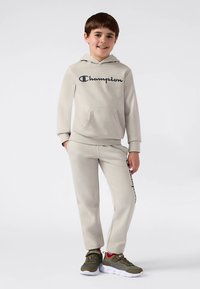 Completo grigio chiaro composto da felpa con cappuccio e pantaloni della tuta, con tasca frontale e logo "Champion". Tessuto strutturato, polsini elasticizzati e sneakers abbinate.
