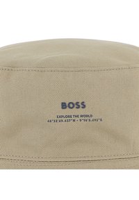 Μπεζ καπέλο τύπου bucket από υφασμάτινο υλικό με υφή, με το όνομα της μάρκας "BOSS" και τη φράση "EXPLORE THE WORLD" σε μπλε ναυτικό χρώμα.