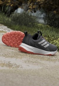 Chaussures de trail Adidas Terrex noires et grises avec semelle extérieure orange, semelle intermédiaire blanche et trois bandes blanches sur un chemin de gravier.
