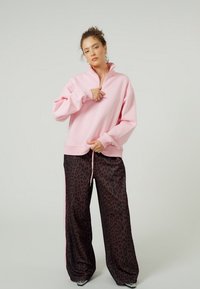 Rozā fliisveida džemperis ar ceturtdaļas rāvējslēdzēja apkakli, kopā ar vaļīgām, brūnā leoparda raksta biksēm ar sānu svītrām.