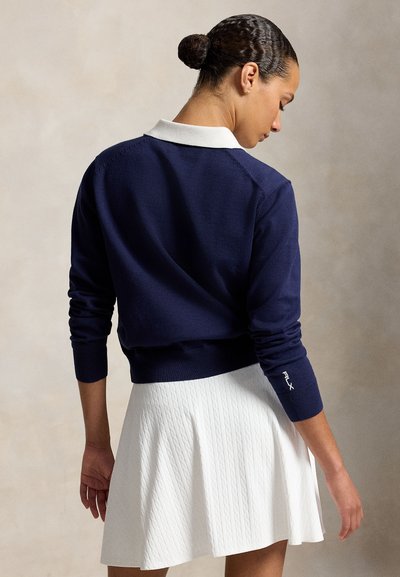Femme avec les cheveux en chignon portant une chemise bleu marine à manches longues avec un col blanc et une jupe blanche texturée, de dos.