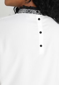 Femme portant une blouse blanche avec trois boutons noirs dans le dos et un col en dentelle noire avec une bordure volantée.