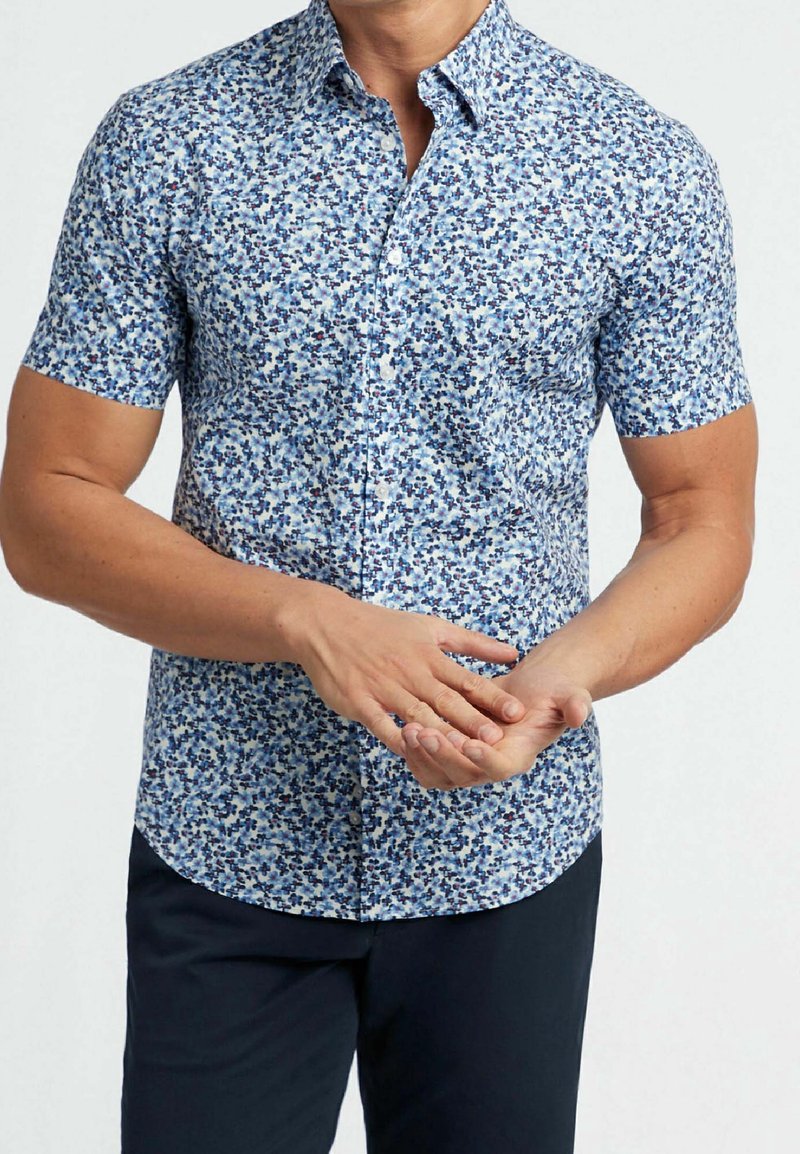 Homme portant une chemise à manches courtes bleue et blanche à motifs floraux avec un pantalon foncé, ajustant une main avec l'autre.