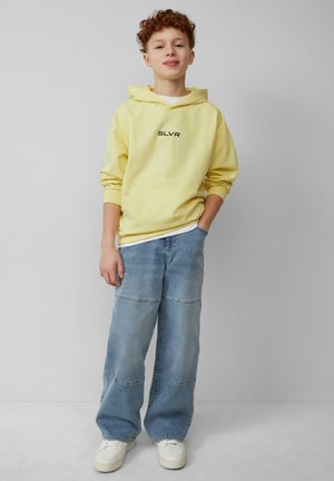 Geel hoodie met de tekst "SLVR", gecombineerd met lichtblauwe wijd uitlopende jeans met patchworkdetails. Witte sneakers maken de outfit compleet.
