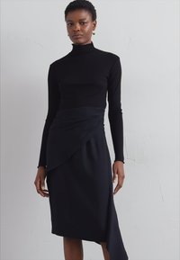 Maglietta nera a maniche lunghe con collo alto abbinata a una gonna al ginocchio con dettaglio drappeggiato e orlo asimmetrico. Tessuto liscio con una silhouette aderente.