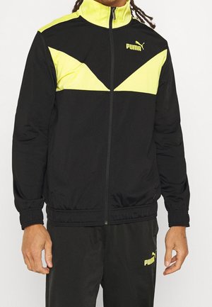Mężczyzna ubrany w czarny dres Puma z żółtymi geometrycznymi wzorami na kurtce i dopasowanych spodniach, stojący na jednolitym tle.