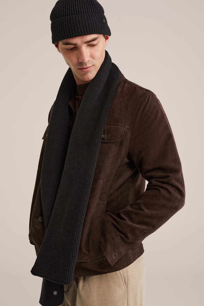 Homme portant une veste en daim marron foncé, une écharpe en tricot noire et un bonnet noir, regardant vers le bas avec une main dans la poche sur fond beige.