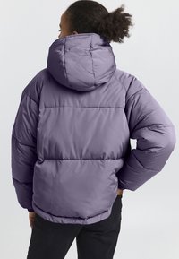Giacca a palloncino viola con cappuccio, caratterizzata da un design trapuntato, polsini elasticizzati e una vestibilità oversize. Texture liscia e tessuto morbido.