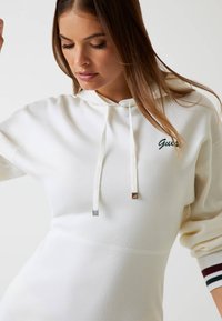 Sudadera blanca de tejido suave que cuenta con un bolsillo canguro frontal, capucha con cordón y puntas plateadas, y logotipo bordado de "Guess" en verde.