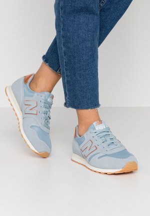 Person iført blå og beige New Balance-sneakers med lyseblå jeans, stående på en hvid gulv mod en ensfarvet baggrund.