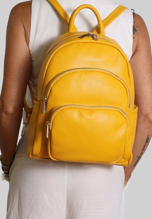 Persoon draagt een mouwloze witte jurk met een gele leren rugzak met meerdere met rits afgesloten vakken.