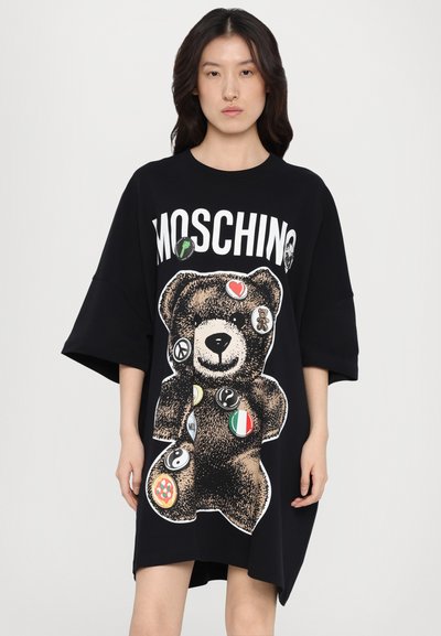 Μαύρο oversized φόρεμα μπλούζας με μεγάλο γραφικό αρκούδας και διάφορες διακριτικές ετικέτες. Κατασκευασμένο από μαλακό ύφασμα, με στρογγυλή λαιμόκοψη και κοντά μανίκια.