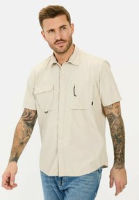 Helles beige Hemd mit kurzen Ärmeln, zwei Brusttaschen, Knopfverschluss und einer subtilen Textur. Verfügt über eine Schlaufe an einer Tasche.