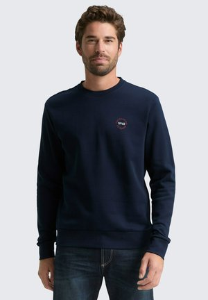 Uomo che indossa una felpa blu navy con un logo rosso sul petto e jeans scuri, in piedi contro uno sfondo grigio chiaro uniforme.