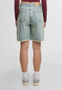 Persona con pantalones cortos de mezclilla azul claro con dobladillo sin terminar, calcetines blancos y zapatillas Adidas grises, de pie contra un fondo blanco liso.