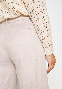 Světlé béžové lněné kalhoty s hladkou texturou, vybavené dvěma zadními kapsami, v kombinaci s krémovou blůzou se vzorem děr.