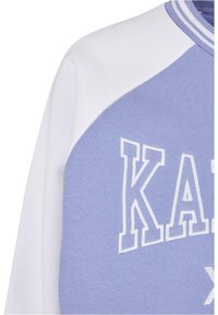 Světle modrá mikina s bílými raglánovými rukávy, na přední straně s vyšívaným nápisem "KAT" v bílé barvě. Měkká látka s žebrovaným límcem.