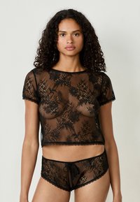 Top corto de encaje negro con mangas cortas y patrones florales, combinado con pantalones cortos de encaje a juego que cuentan con cordón y bordes en forma de ondas.