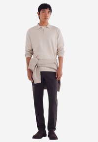 Beiger Reißverschluss-Pullover über einem grauen Rundhalshemd, kombiniert mit dunkelgrauen Hosen und braunen Schuhen. Der Pullover hat gerippte Bündchen und einen gerippten Saum.