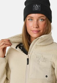 Giacca in fleece beige con chiusura a zip sul fronte e tasca laterale, con dettagli del logo. Berretto in maglia nera con patch del logo. La texture del fleece è visibile.
