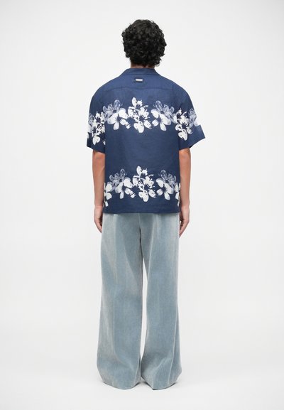 Chemise à manches courtes marine avec un imprimé floral blanc, portée avec un pantalon large bleu clair, mettant en valeur une coupe décontractée et un tissu texturé.