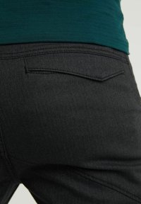 Close-up van zwarte gestructureerde broek met achterzakflap, gedragen met een donkergroen bovenstuk.